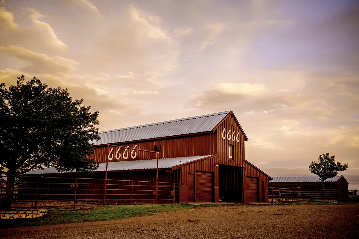 Red barn displaying the 6666 logo at Four Sixes Ranch, surrounded by corrals beneath a Texas sunset sky.