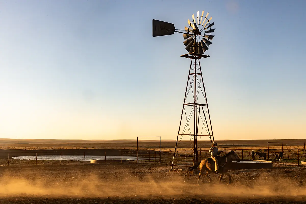 A cowboy on horseback crossing open rangeland by a windmill at dusk on Four Sixes Ranch.